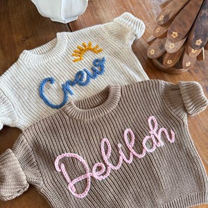 K&ouml;nnte beinhalten: Zwei Strickpullover. Einer ist cremefarben mit dem Wort "Crew" in Blau und einem gelben Sonnendesign. Der andere ist braun mit dem Namen "Delilah" in Rosa. Beide haben gerippte B&uuml;ndchen und Rundhalsausschnitte.