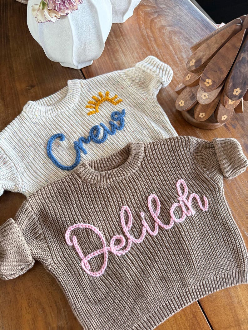 K&ouml;nnte beinhalten: Zwei Strickpullover. Einer ist cremefarben mit dem Wort "Crew" in Blau und einem gelben Sonnendesign. Der andere ist braun mit dem Namen "Delilah" in Rosa. Beide haben gerippte B&uuml;ndchen und Rundhalsausschnitte.