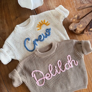 K&ouml;nnte beinhalten: Zwei Baby-Pullover. Einer ist cremefarben mit dem Wort "Crew" in Blau mit einer gelben Sonne gestickt. Der andere ist braun mit dem Namen "Delilah" in Rosa gestickt. Beide Pullover haben gerippte B&uuml;ndchen und einen runden Halsausschnitt.