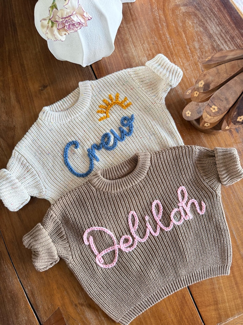 K&ouml;nnte beinhalten: Zwei Baby-Pullover. Einer ist cremefarben mit dem Wort "Crew" in Blau mit einer gelben Sonne gestickt. Der andere ist braun mit dem Namen "Delilah" in Rosa gestickt. Beide Pullover haben gerippte B&uuml;ndchen und einen runden Halsausschnitt.