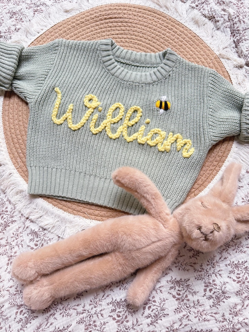 K&ouml;nnte beinhalten: Hellgr&uuml;ner Strickpullover mit dem Namen "William" in gelber Schrift und einer kleinen Bienenapplikation. Ein hellbraunes Pl&uuml;schkaninchen liegt darunter auf einer floral gemusterten Oberfl&auml;che. Der Pullover hat lange &Auml;rmel.