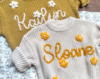 May include: Two knitted sweaters, one mustard yellow and one beige, are displayed. The yellow sweater has the name "Kailyn" embroidered in white, with white flower accents. The beige sweater has the name "Sloane" embroidered in yellow, with yellow flower accents.