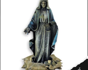 May include: A weathered statue of a robed figure with a hood, standing on sandbags. The statue has a blue and gray color scheme, with the figure's arms outstretched. The base of the statue is surrounded by sandbags. The statue is a religious item.
