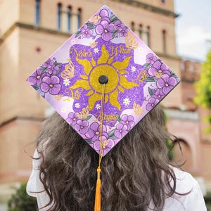 May include: A graduation cap with a purple and gold design. The cap features a sun design with the text "Now's When Life Begins" and is decorated with purple flowers and butterflies. An orange tassel hangs from the cap.