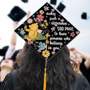 May include: A black graduation cap with a Winnie the Pooh and Piglet design, surrounded by colorful flowers. The cap features the quote, "It makes such a difference said Pooh, to have someone who believes in you."
