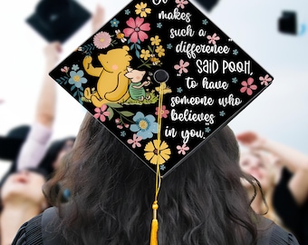 May include: A black graduation cap with a Winnie the Pooh and Piglet design, surrounded by colorful flowers. The cap features the quote, "It makes such a difference said Pooh, to have someone who believes in you."