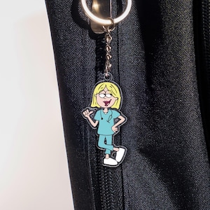 May include: A cartoon keychain featuring a female nurse with blonde hair, glasses, and a stethoscope. She is wearing blue scrubs and white shoes. The keychain is attached to a silver keyring and chain, hanging against a black background.