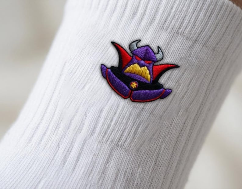 Op de afbeelding: Een witte geribbelde sok met een geborduurde patch van een paars, gehoornd personage met rode accenten, gele tanden en een klein ster-embleem op de borst. Het personage heeft een dreigende uitdrukking met smalle rode ogen en grijze hoorns.