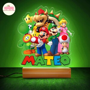 Peut inclure: Lampe de chevet en acrylique color&eacute; repr&eacute;sentant les personnages de Super Mario Bros. Mario, Luigi, Peach et d'autres personnages sont pr&eacute;sents, avec le nom "MATEO" en lettres color&eacute;es. La base est en bois.