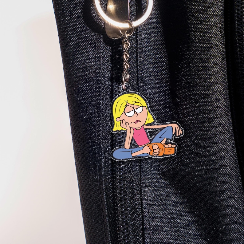 May include: A keychain featuring a cartoon character with blonde hair, wearing a pink top, blue jeans, and orange sandals. The character is sitting with their chin resting on their hand. The keychain is attached to a silver ring and chain.