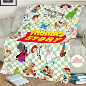 May include: A plush blanket featuring a green and white checkered pattern with various Toy Story characters. The blanket has the text "THOMAS STORY" in large, colorful letters. The blanket is draped over a sofa.