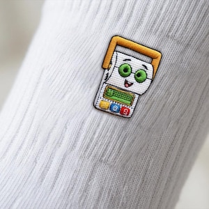Op de afbeelding: Een witte geribbelde sok met een geborduurde patch van een stripfiguur die lijkt op een wc-rol met een geel handvat, groene ogen en een rekenmachine-achtige interface met een gele, blauwe en rode knop aan de onderkant.