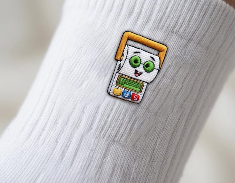 Op de afbeelding: Een witte geribbelde sok met een geborduurde patch van een stripfiguur die lijkt op een wc-rol met een geel handvat, groene ogen en een rekenmachine-achtige interface met een gele, blauwe en rode knop aan de onderkant.