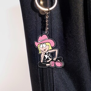 May include: A pink and black cartoon cowgirl keychain. The keychain features a cartoon cowgirl wearing a pink cowboy hat, a black and white cow print top, and black pants. The keychain is attached to a silver keyring.