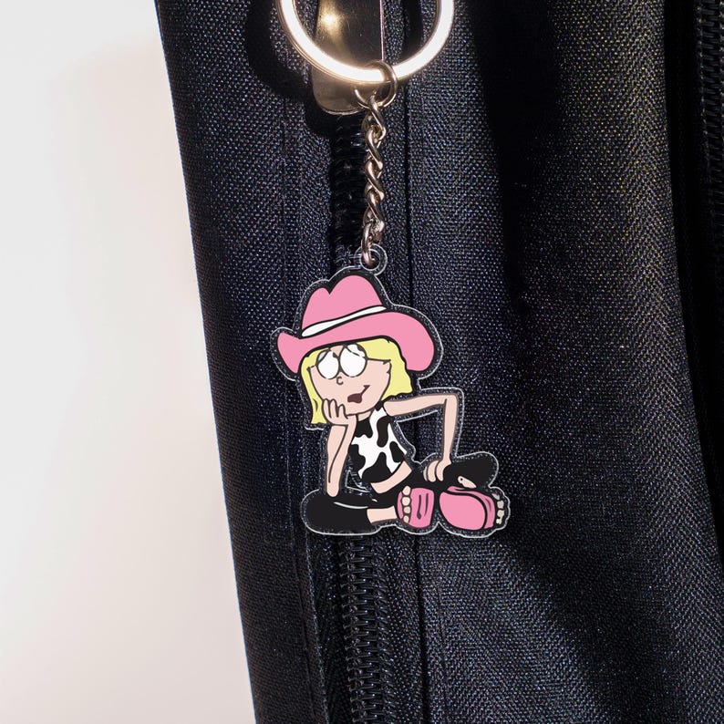 May include: A pink and black cartoon cowgirl keychain. The keychain features a cartoon cowgirl wearing a pink cowboy hat, a black and white cow print top, and black pants. The keychain is attached to a silver keyring.