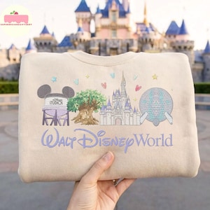 May include: Beige sweatshirt with embroidered Walt Disney World design. Features Mickey Mouse ears, a tree, a castle, and a sphere. The text "Walt Disney World" is embroidered in a cursive font.