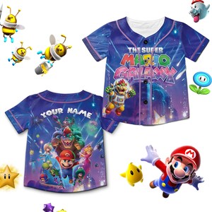 Puede incluir: Una camiseta de b&eacute;isbol azul con personajes y texto del videojuego "The Super Mario Galaxy". La parte delantera de la camiseta muestra el t&iacute;tulo del juego y Bowser. La parte trasera tiene el texto "YOUR NAME" encima de un gr&aacute;fico de Mario y otros personajes.