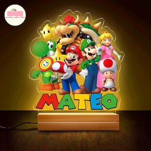 Peut inclure: Lampe de chevet LED color&eacute;e repr&eacute;sentant des personnages de Super Mario, dont Mario, Luigi et la Princesse Peach. Le nom "MATEO" est affich&eacute; en grandes lettres color&eacute;es. La lampe est pos&eacute;e sur une base en bois.