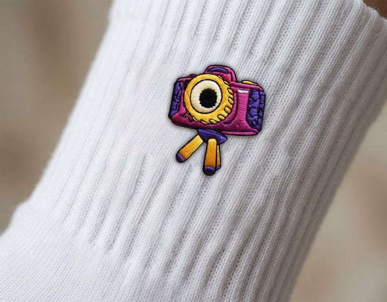 Op de afbeelding: Een witte geribbelde sok met een geborduurde patch van een camera op een statief. De camera is magenta en paars met een opvallende gele lens, terwijl de statiefpoten geel zijn met paarse uiteinden, wat een kleurrijk grafisch detail vormt.