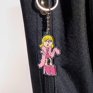 May include: A pink and yellow cartoon keychain featuring a woman in a pink dress, pink platform shoes, and a pink boa. The keychain is attached to a silver keyring and chain. The background is a black fabric.