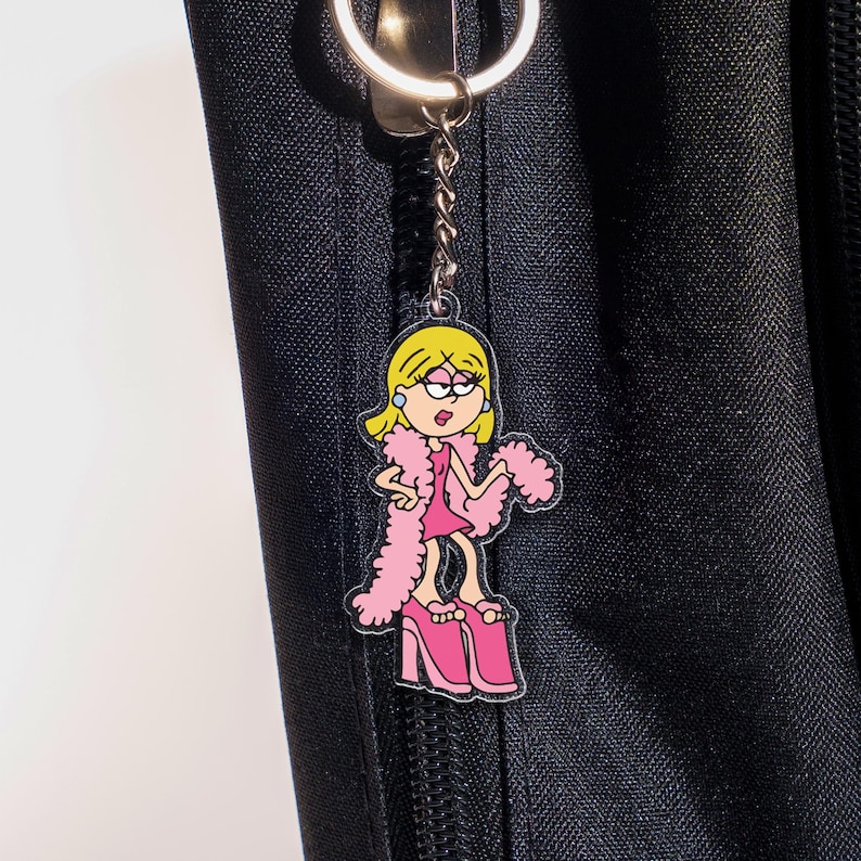 May include: A pink and yellow cartoon keychain featuring a woman in a pink dress, pink platform shoes, and a pink boa. The keychain is attached to a silver keyring and chain. The background is a black fabric.