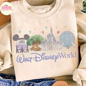 May include: A cream-coloured sweatshirt with a Walt Disney World design. The graphic includes the Disney Studios water tower, the Tree of Life, Cinderella Castle, and Spaceship Earth, with the words "Walt Disney World" in a script font. Small hearts and stars are scattered above the design.