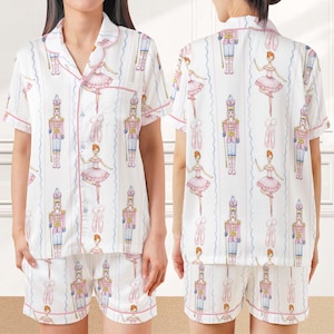 May include: White pajama set with a repeating pattern of ballerinas, nutcrackers, and ballet slippers. The short-sleeved top and shorts are trimmed in pink. The fabric has vertical blue and white striped accents.