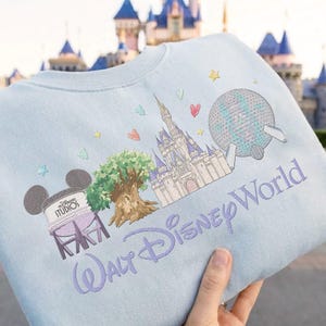 May include: Pale blue sweatshirt with an embroidered design of Walt Disney World icons. The design includes Cinderella's castle, Spaceship Earth, the Tree of Life, and the words "Walt Disney World" in a purple script. The sweatshirt is held up by a hand.