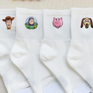Op de afbeelding: Vier witte crew-sokken met geborduurde karakterhoofden uit Toy Story. De eerste sok toont een cowboy, de tweede een space ranger, de derde een roze varken en de vierde een bruine en beige teckel met een veerachtig lichaam.