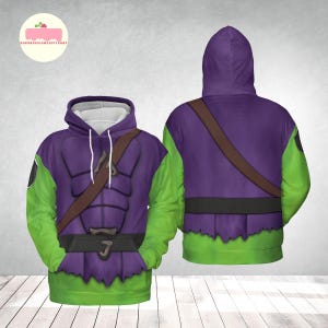 May include: A hooded sweatshirt featuring a purple torso with a brown strap and green sleeves. The design resembles a costume with a jagged hemline and a black emblem on the left sleeve. The hoodie has a drawstring and a white interior.