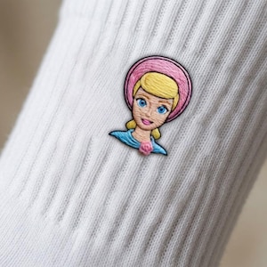 Op de afbeelding: Een witte geribbelde sok met een geborduurde patch van een personage met blond haar, blauwe ogen en een roze hoed. Het personage draagt een blauwe top met een klein roze roosdetail bij de halslijn, uitgevoerd in gedetailleerd borduurwerk.