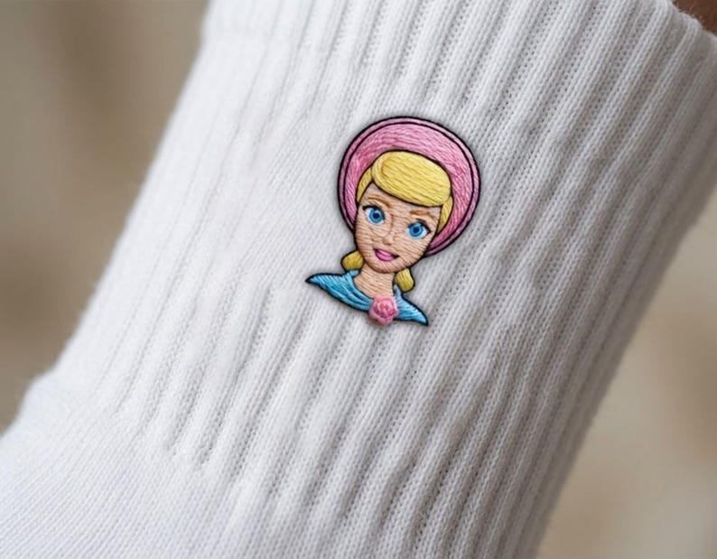 Op de afbeelding: Een witte geribbelde sok met een geborduurde patch van een personage met blond haar, blauwe ogen en een roze hoed. Het personage draagt een blauwe top met een klein roze roosdetail bij de halslijn, uitgevoerd in gedetailleerd borduurwerk.