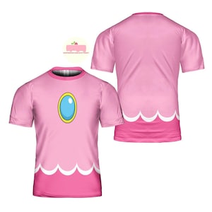 May include: Pink t-shirt featuring a design inspired by Princess Peach from the Super Mario video game series. The shirt has a large blue and gold jewel graphic on the front, with a white scalloped hemline. The back of the shirt is plain pink.