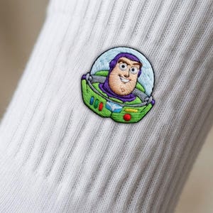 Op de afbeelding: Een witte geribbelde sok met een gedetailleerde geborduurde patch van het personage Buzz Lightyear. Het borduurwerk toont het personage in een groen en paars ruimtepak met een doorzichtige helmkoepel in levendige kleuren.