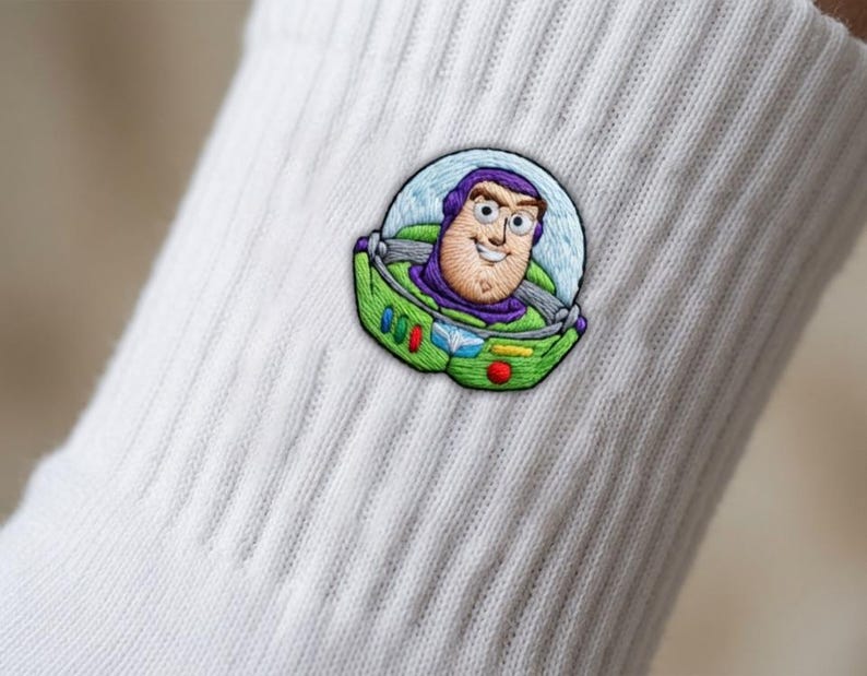 Op de afbeelding: Een witte geribbelde sok met een gedetailleerde geborduurde patch van het personage Buzz Lightyear. Het borduurwerk toont het personage in een groen en paars ruimtepak met een doorzichtige helmkoepel in levendige kleuren.