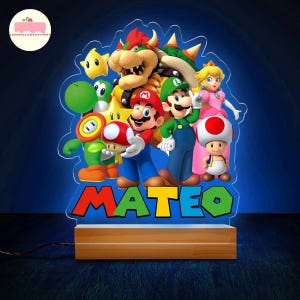 Peut inclure: Veilleuse en acrylique color&eacute;e repr&eacute;sentant les personnages de Super Mario, dont Mario, Luigi et la Princesse Peach. Le nom "MATEO" est affich&eacute; en bas. La lampe est pos&eacute;e sur un socle en bois.