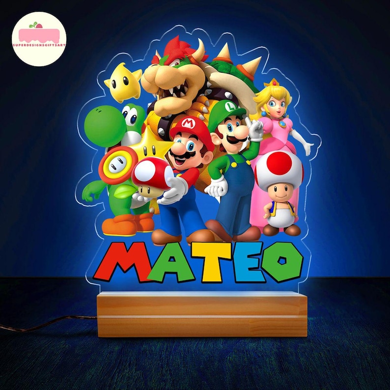 Peut inclure: Veilleuse en acrylique color&eacute;e repr&eacute;sentant les personnages de Super Mario, dont Mario, Luigi et la Princesse Peach. Le nom "MATEO" est affich&eacute; en bas. La lampe est pos&eacute;e sur un socle en bois.