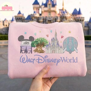 May include: A pale pink sweatshirt with embroidered Disney World imagery. The design includes the Hollywood Studios water tower, the Tree of Life, Cinderella's Castle, and Spaceship Earth. The words "Walt Disney World" are embroidered in light blue.