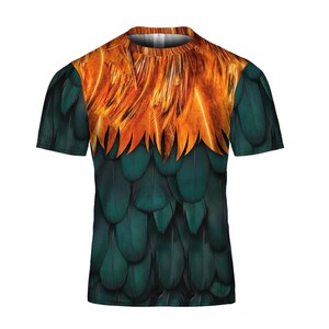 Puede incluir: Una camiseta de manga corta con un llamativo dise&ntilde;o de plumas. La parte superior muestra plumas naranjas y amarillas, que se funden en plumas verde oscuro. La camiseta tiene cuello redondo.