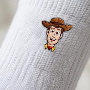 Op de afbeelding: Een witte geribbelde sok met een geborduurde patch van een lachend cowboy-personage met een bruine hoed, een geel overhemd en een rode halsdoek. Het personage heeft een lichte huid, bruin haar en grote ogen, gecentreerd op de stof.