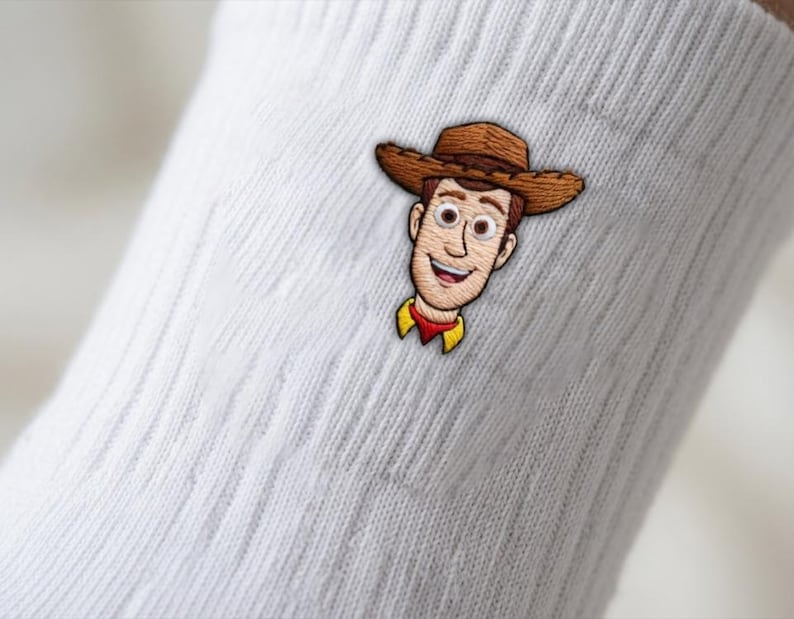 Op de afbeelding: Een witte geribbelde sok met een geborduurde patch van een lachend cowboy-personage met een bruine hoed, een geel overhemd en een rode halsdoek. Het personage heeft een lichte huid, bruin haar en grote ogen, gecentreerd op de stof.