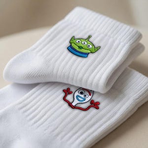 Op de afbeelding: Een paar witte geribbelde sokken met geborduurde karakterontwerpen. De ene sok toont een groene alien met drie ogen en een blauwe kraag, terwijl de andere sok een wit vork-figuurtje met rode armen en blauwe ogen laat zien.