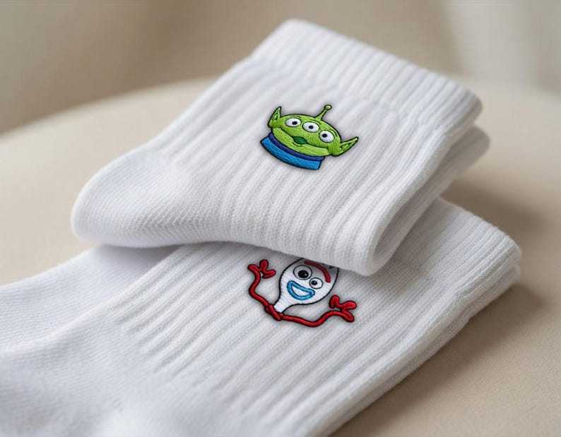 Op de afbeelding: Een paar witte geribbelde sokken met geborduurde karakterontwerpen. De ene sok toont een groene alien met drie ogen en een blauwe kraag, terwijl de andere sok een wit vork-figuurtje met rode armen en blauwe ogen laat zien.