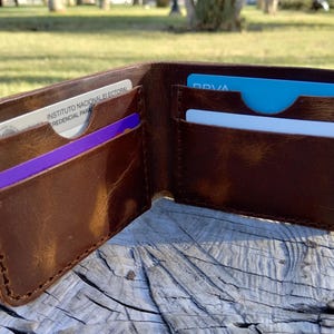 Könnte beinhalten: Offene braune Lederbrieftasche mit mehreren Kartenfächern. Die Brieftasche enthält mehrere Karten in verschiedenen Farben, darunter Blau, Lila und Weiß. Die Brieftasche ist geöffnet und zeigt das Innere und die Nähte.