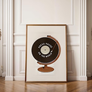 May include: A framed print featuring a brown globe design with a record-like appearance. The text "PRESS PAUSE ON THE WORLD" is printed on the record. The globe is set against a cream-colored background and framed in a wooden frame. The print is displayed on a wooden floor.