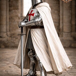 May include: A full suit of medieval knight armor, including a helmet, chest plate with a red cross, chainmail, and leg protection. The knight wears a white cape and holds a long sword. The image is set in a stone building with a Gothic Medieval Mart logo.