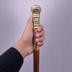 Puede incluir: Bastón decorativo con un mango dorado detallado y un eje de madera marrón oscuro. El mango presenta tallas ornamentadas y se sostiene en una mano. El bastón mide aproximadamente 91 cm de largo.