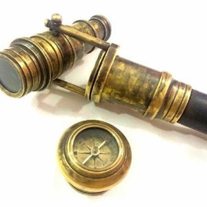 Puede incluir: Telescopio antiguo de latón con un mango de madera oscura y una pequeña brújula de latón. El telescopio tiene múltiples secciones y una lente. La brújula tiene una cubierta de vidrio y una aguja que apunta hacia el norte.
