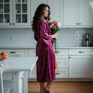 May include: A woman in a magenta velvet robe holding a bouquet of flowers in a bright white kitchen. The kitchen has white cabinets, a white countertop, and a stainless steel mixer. A bowl of fruit sits on the table.