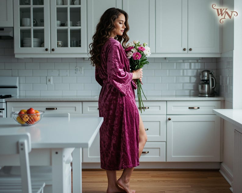 May include: A woman in a magenta velvet robe holding a bouquet of flowers in a bright white kitchen. The kitchen has white cabinets, a white countertop, and a stainless steel mixer. A bowl of fruit sits on the table.